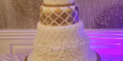 A four-tier white wedding cake featuring gold lattice patterns, pearl accents, and a floral topper, displayed on a gold stand.