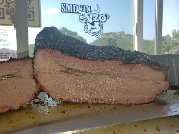 Sliced smoked brisket with a dark bark and pink smoke ring, displayed in front of a Smokin Zo's logo.