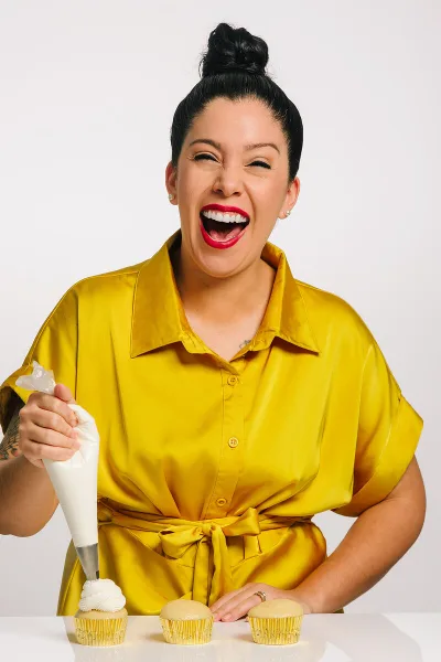 A smiling woman in a yellow shirt piping white frosting onto a cupcake.