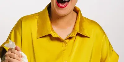 A smiling woman in a yellow shirt piping white frosting onto a cupcake.