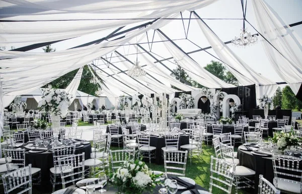 A large white event tent interior featuring draped fabric, hanging chandeliers, and rows of round tables set with white chairs and floral arrangements.