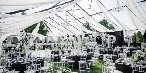 A large white event tent interior featuring draped fabric, hanging chandeliers, and rows of round tables set with white chairs and floral arrangements.
