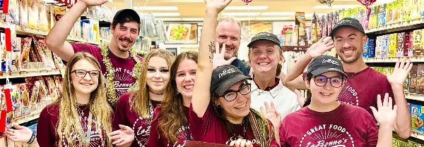 A group of smiling LaBonne's Markets employees wearing maroon branded shirts and caps, posing together in a grocery store aisle.