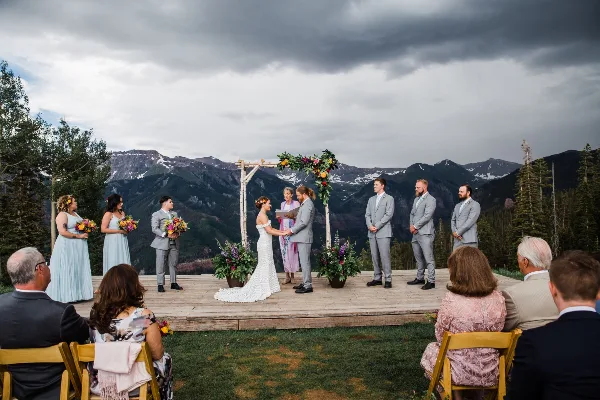 A wedding ceremony taking place on a wooden stage with a mountain view, featuring a bride, groom, and officiant.