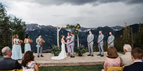 A wedding ceremony taking place on a wooden stage with a mountain view, featuring a bride, groom, and officiant.