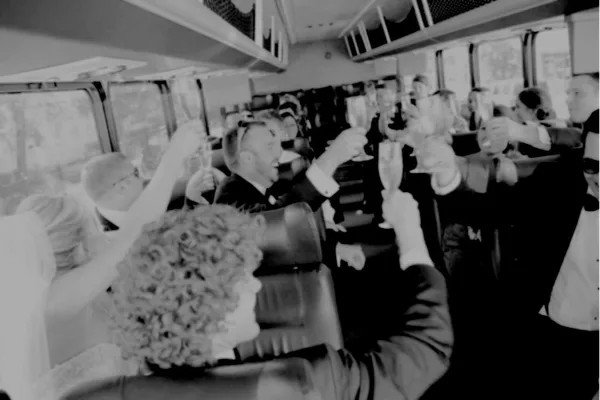 A black and white photo of wedding guests and a bride toasting with champagne inside a shuttle bus.