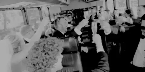 A black and white photo of wedding guests and a bride toasting with champagne inside a shuttle bus.