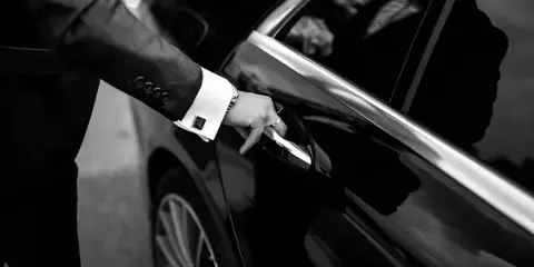 A professional chauffeur in a suit opening the door of a black luxury sedan.