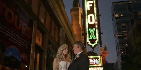 A bride and groom standing together in front of the illuminated Fox Theatre sign at dusk.