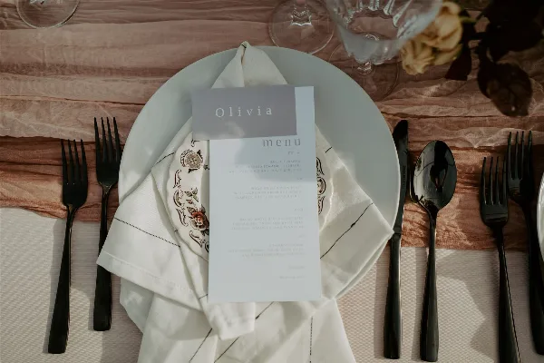 Styled wedding table setting with white plate, embroidered napkin, place card reading Olivia, menu card, black flatware, and glassware on draped linens.