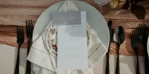 Styled wedding table setting with white plate, embroidered napkin, place card reading Olivia, menu card, black flatware, and glassware on draped linens.