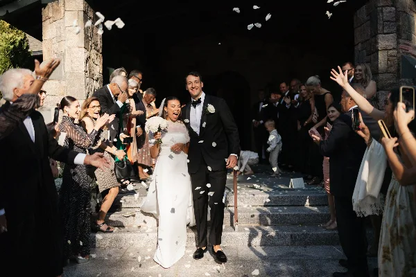 A bride and groom walk down stone steps surrounded by guests throwing white petals during a wedding exit.