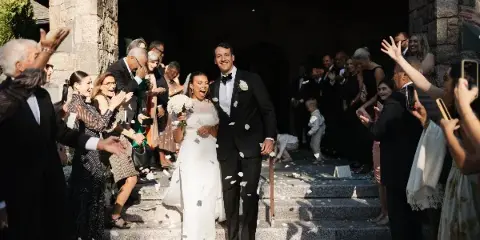 A bride and groom walk down stone steps surrounded by guests throwing white petals during a wedding exit.