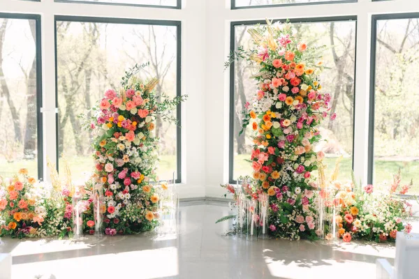 A wedding ceremony installation featuring two tall, colorful floral pillars with roses, dahlias, and greenery, surrounded by ground arrangements and candles.