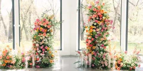 A wedding ceremony installation featuring two tall, colorful floral pillars with roses, dahlias, and greenery, surrounded by ground arrangements and candles.