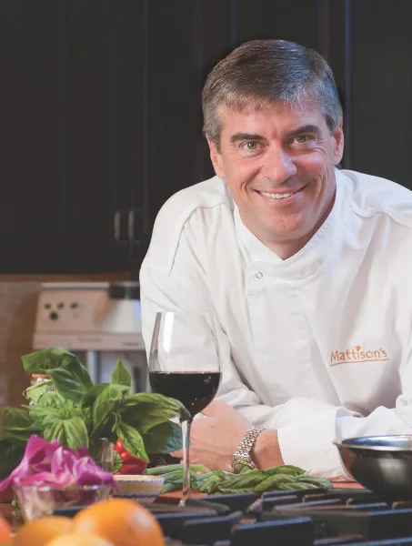 A smiling chef in a white uniform stands in a kitchen with fresh vegetables and a glass of wine.