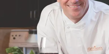 A smiling chef in a white uniform stands in a kitchen with fresh vegetables and a glass of wine.