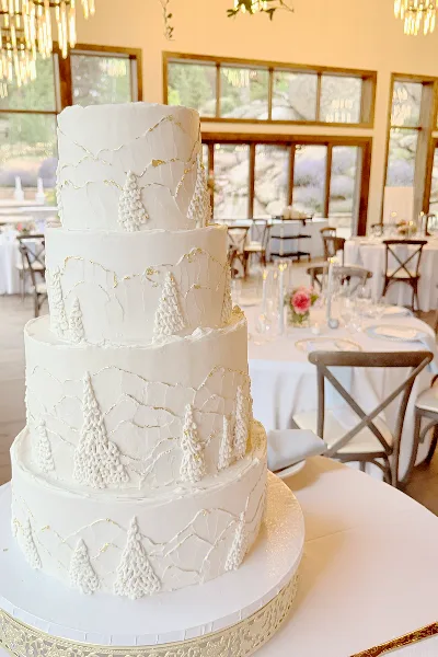 A four-tier white wedding cake decorated with piped mountain ranges and pine trees, displayed on a table in a banquet hall.
