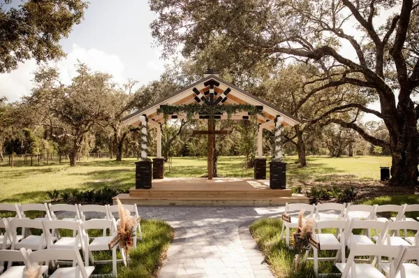 Outdoor wedding ceremony setup with white chairs, a paved aisle, and a wooden altar with a cross under oak trees.