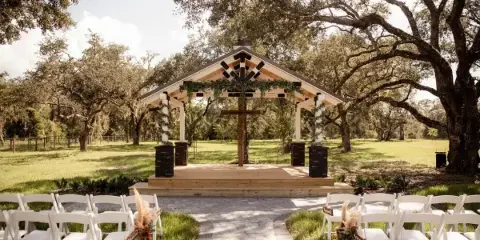 Outdoor wedding ceremony setup with white chairs, a paved aisle, and a wooden altar with a cross under oak trees.