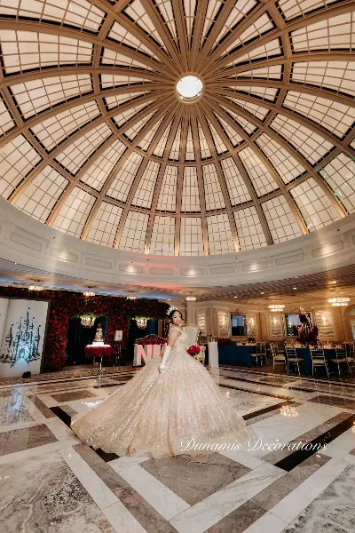 A bride in a white gown standing in a grand ballroom with a domed ceiling, surrounded by red floral decorations and set dining tables.