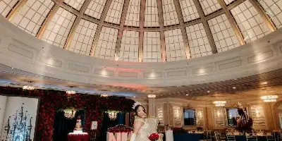 A bride in a white gown standing in a grand ballroom with a domed ceiling, surrounded by red floral decorations and set dining tables.
