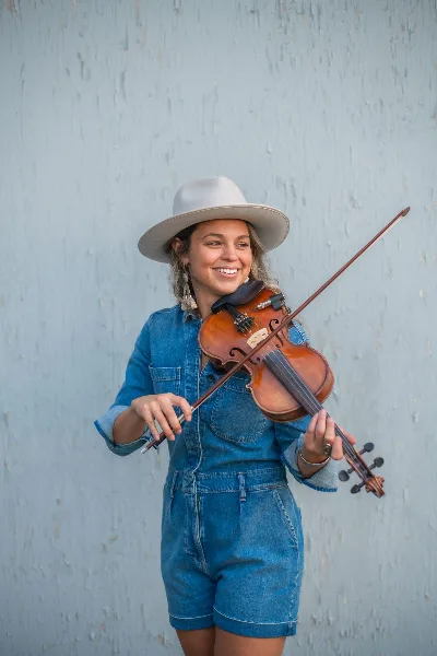 A smiling woman in a denim outfit and hat holding a fiddle and bow against a light blue wall.
