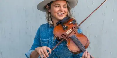 A smiling woman in a denim outfit and hat holding a fiddle and bow against a light blue wall.