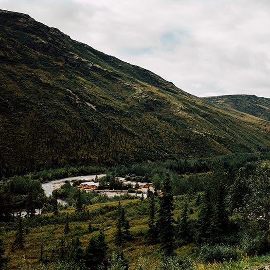 A lodge complex with multiple buildings situated in a valley surrounded by forested mountains and evergreen trees.