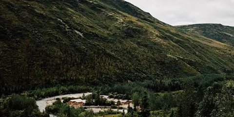 A lodge complex with multiple buildings situated in a valley surrounded by forested mountains and evergreen trees.