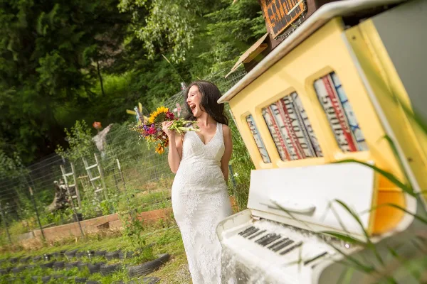 A bride in a white lace wedding dress holding a colorful bouquet stands outdoors near a yellow vintage prop and green foliage.