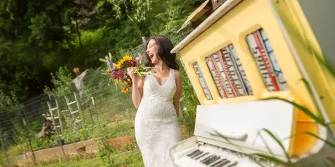 A bride in a white lace wedding dress holding a colorful bouquet stands outdoors near a yellow vintage prop and green foliage.