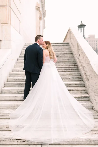 A bride and groom standing on stone steps, the groom kissing the bride's forehead while she looks down, wearing a long white wedding gown.