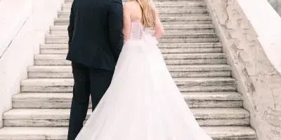 A bride and groom standing on stone steps, the groom kissing the bride's forehead while she looks down, wearing a long white wedding gown.