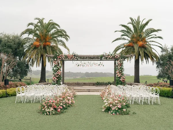 An outdoor wedding ceremony setup featuring a floral arch, white chairs, and pink rose arrangements on a green lawn.