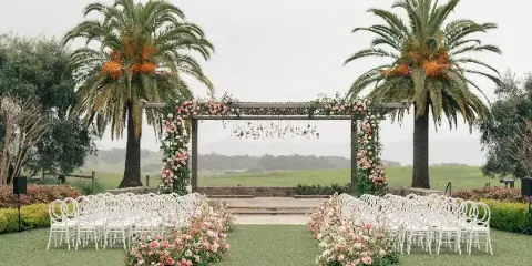 An outdoor wedding ceremony setup featuring a floral arch, white chairs, and pink rose arrangements on a green lawn.