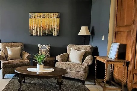 Interior lounge area of a lodge with upholstered armchairs, a coffee table, lamp, and wall art.