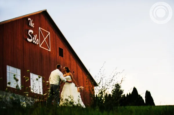 A bride and groom standing in front of a red barn with 'The Silo' signage.