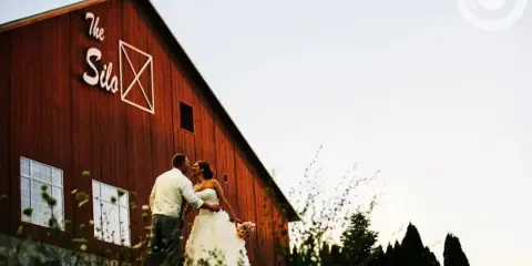 A bride and groom standing in front of a red barn with 'The Silo' signage.