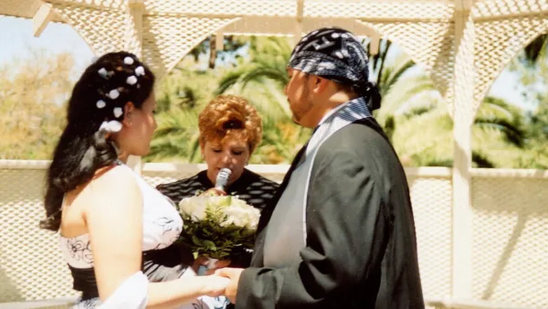 A wedding officiant performing a ceremony for a bride and groom outdoors under a white trellis.