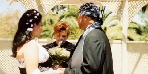 A wedding officiant performing a ceremony for a bride and groom outdoors under a white trellis.