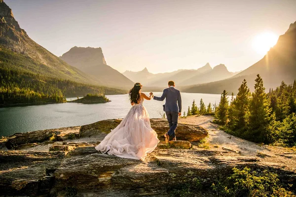 A bride and groom holding hands on a rocky outcrop overlooking a mountain lake at sunset.