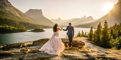 A bride and groom holding hands on a rocky outcrop overlooking a mountain lake at sunset.