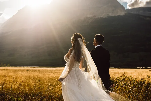 A bride and groom standing in a golden field with a mountain backdrop during sunset.