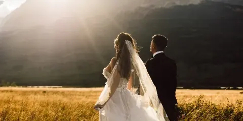A bride and groom standing in a golden field with a mountain backdrop during sunset.