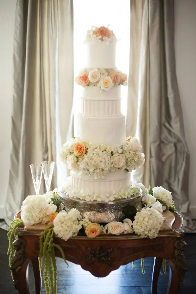 A five-tier white wedding cake decorated with white and peach roses and hydrangeas, displayed on an antique wooden table with champagne flutes.