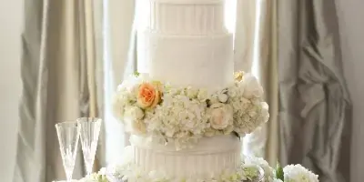 A five-tier white wedding cake decorated with white and peach roses and hydrangeas, displayed on an antique wooden table with champagne flutes.