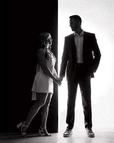 Black and white studio portrait of a couple holding hands, standing against a high-contrast background.