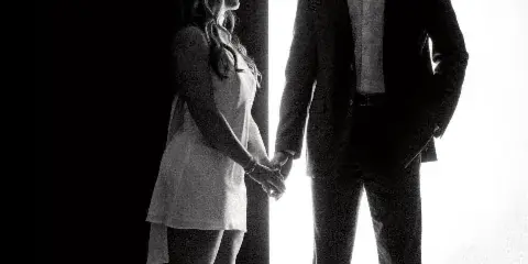 Black and white studio portrait of a couple holding hands, standing against a high-contrast background.