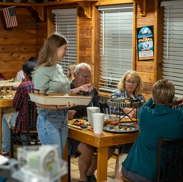 A server carrying a tray in a wood-paneled restaurant dining room with seated guests and food on the table.